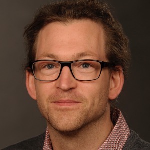 Porträt Foto eines Mannes. Dreitagebart, schwarze Brille mit dicken Rändern. Lange Haare zum Zopf gebunden.